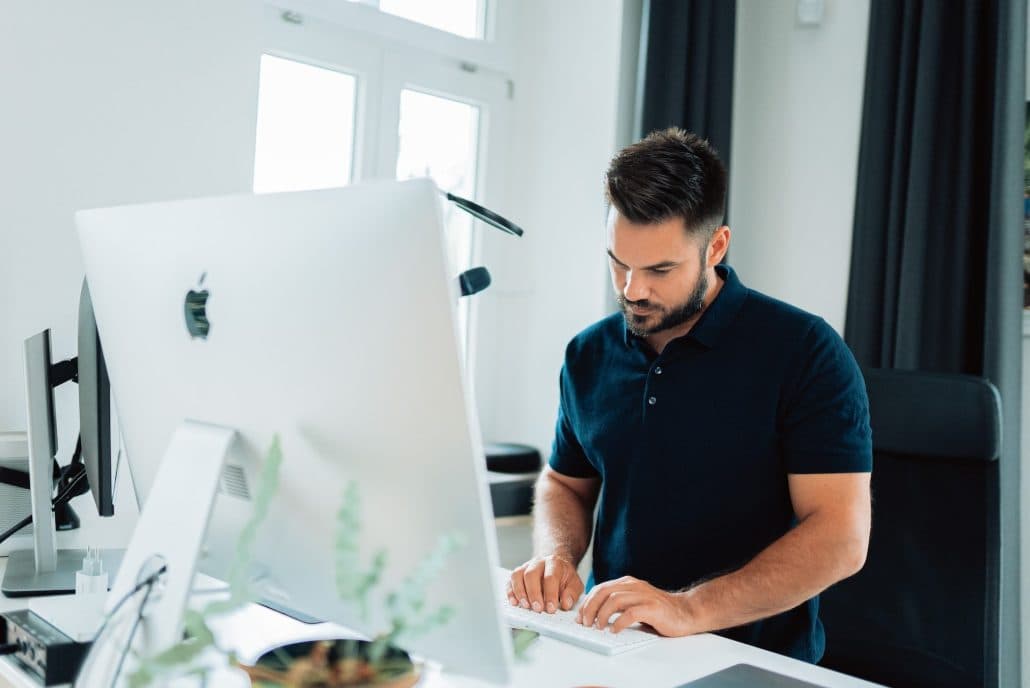 Man in a navy polo typing on a keyboard at an iMac in a bright office