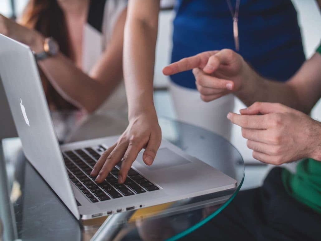 Hands pointing at a laptop on a glass table while colleagues look on