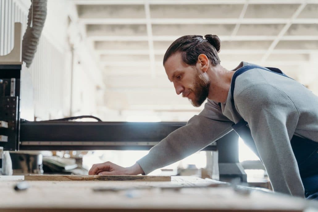 Man with a beard leaning over a wooden board on a woodworking machine in a workshop